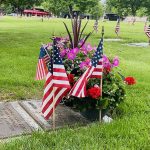 Pink and red flowers and small US flags on a gravestone in a cemetery. Additional headstones and US flags are seen in the green grass background.