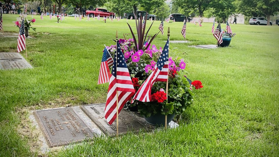Pink and red flowers and small US flags on a gravestone in a cemetery. Additional headstones and US flags are seen in the green grass background.