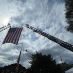 Two firetruck ladders raised high with a large US Flag hanging between them, thin clouds and sunlight fill the background.