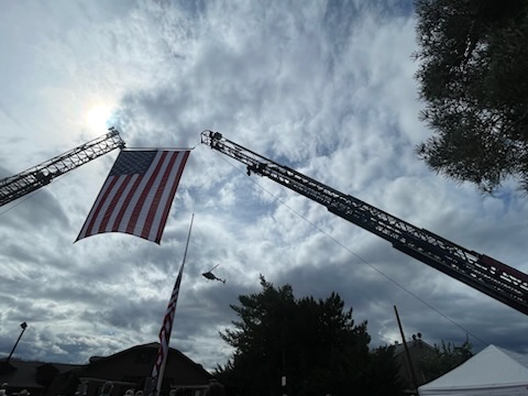 Two firetruck ladders raised high with a large US Flag hanging between them, thin clouds and sunlight fill the background.