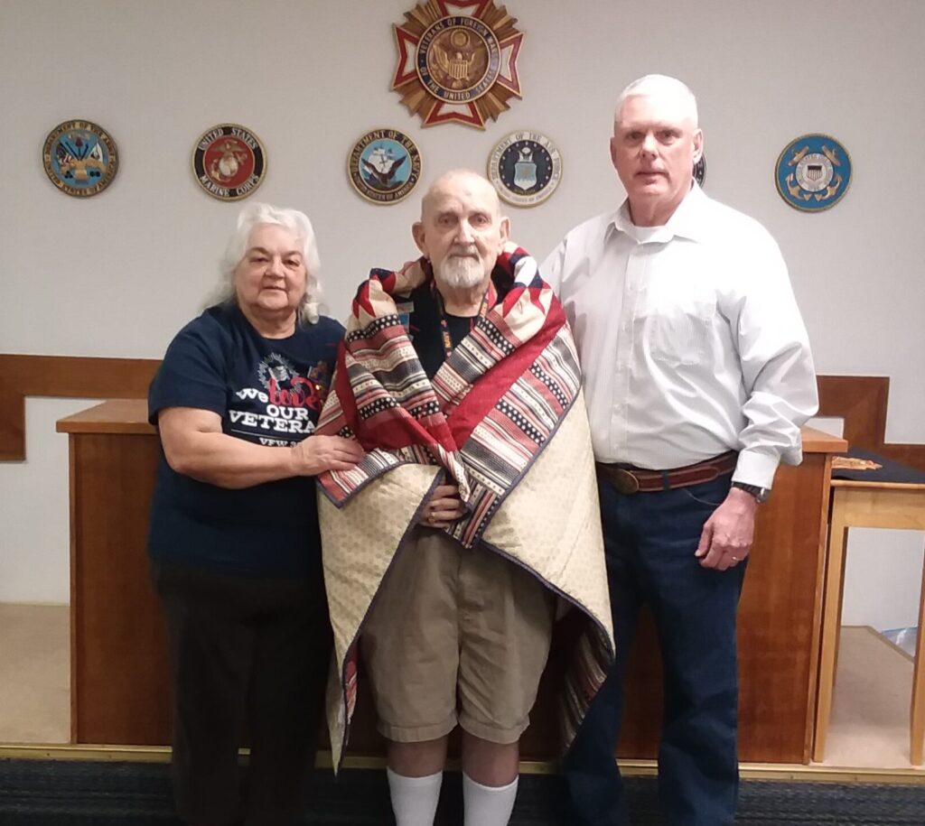 Woman and man standing to either side of a standing veteran wrapped in a patriotic quilt.