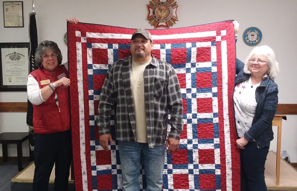 Two women holding up a patriotic quilt behind a standing veteran.